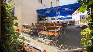 Ein Außenbereich mit Tischen und Stühlen unter blauen Sonnenschirmen, ein Gebäude mit dem Schild "Hofbräu München" und ein Garten mit Pflanzen.