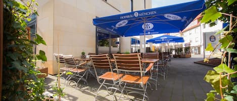 Un espacio al aire libre con mesas y sillas bajo sombrillas azules, un edificio con un letrero que dice "Hofbräu München", y un jardín con plantas.