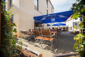 Uma área externa com mesas e cadeiras sob guarda-sóis azuis, um edifício com a placa "Hofbräu München" e um jardim com plantas.