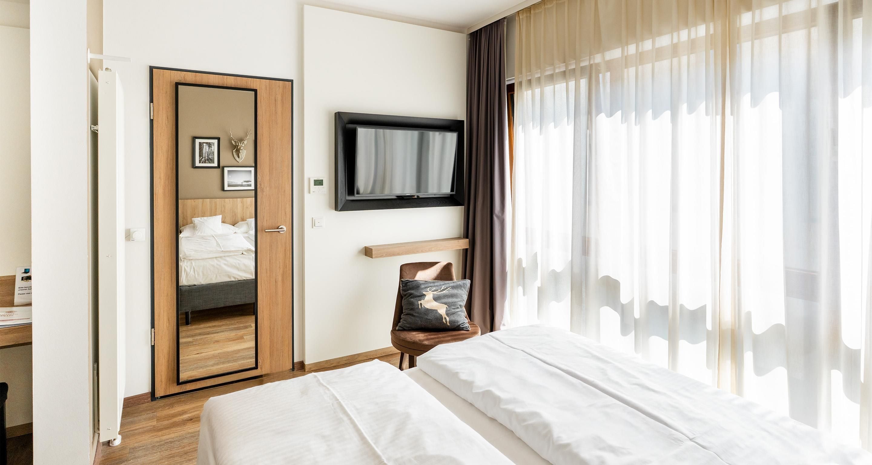 Une chambre d’hôtel avec un lit, un téléviseur fixé au mur, une armoire en bois et une fenêtre avec des rideaux légers.