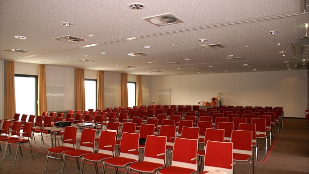Ein Konferenzraum mit Reihen roter Stühle, einer Bühne mit Rednerpult und einer Decke mit eingelassenen Leuchten.