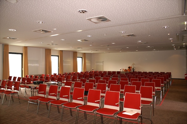 Uma sala de conferências com fileiras de cadeiras vermelhas, um palco com um pódio e um teto com iluminação embutida.