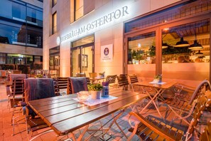 Un área de asientos al aire libre con mesas y sillas, un edificio con un letrero que dice 'HOFBRÜD AM OSTERTOR', y una vista del interior a través de las ventanas.