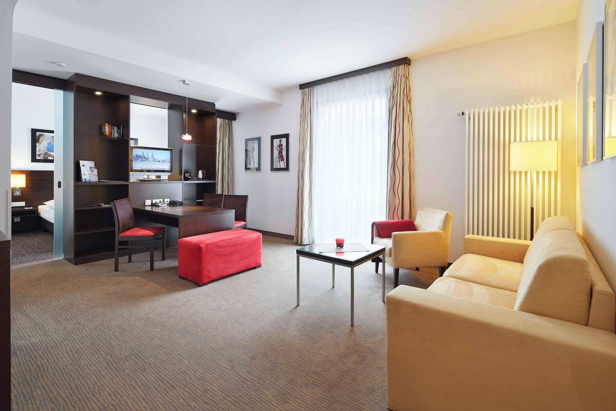 Une chambre d’hôtel avec un canapé, un pouf rouge, une petite table, un téléviseur, un bureau et une bibliothèque.
