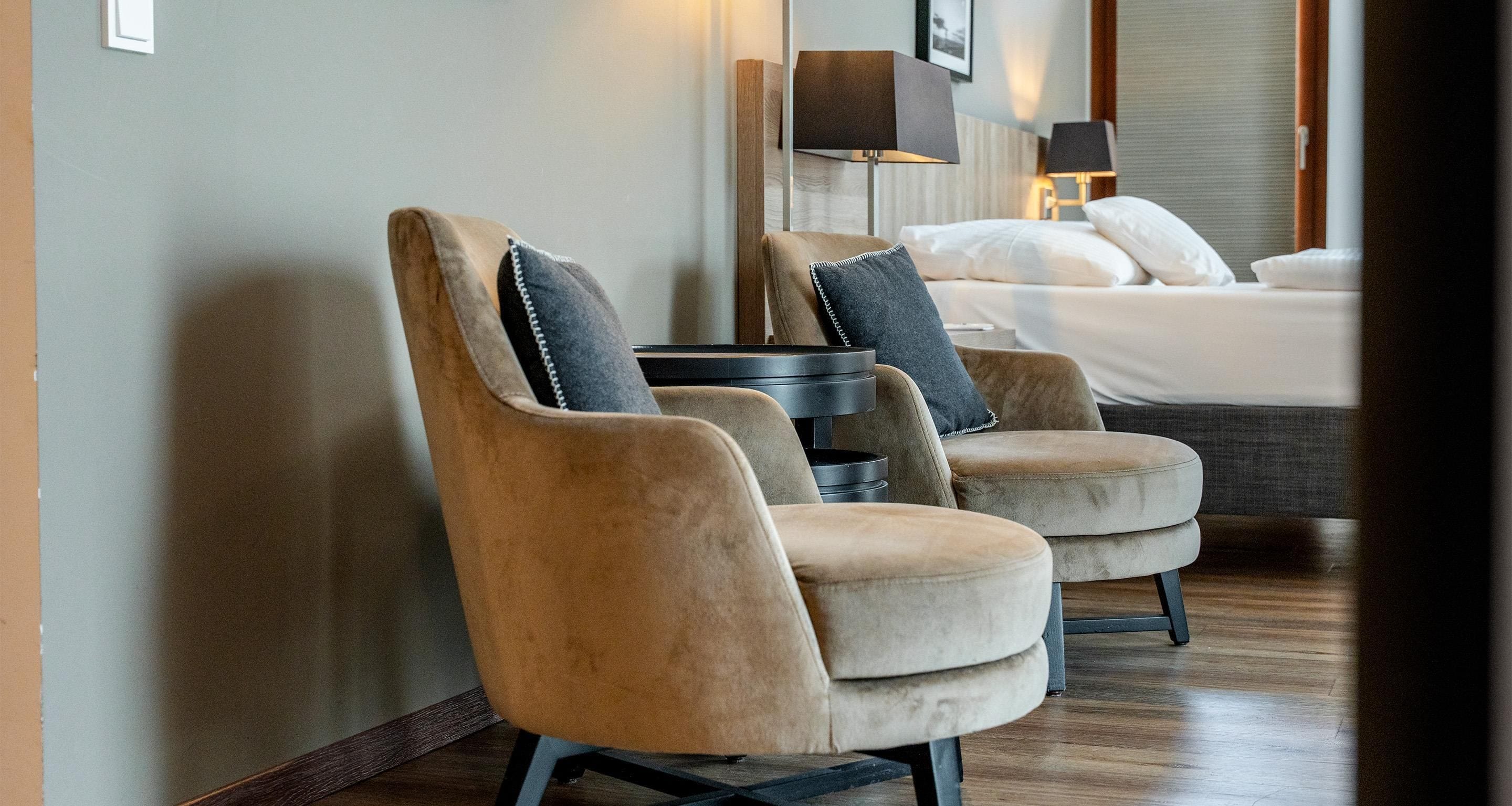 Ein Hotelzimmer mit zwei bequemen Sesseln, einem Bett mit weißer Bettwäsche und einem Nachttisch mit Lampe.