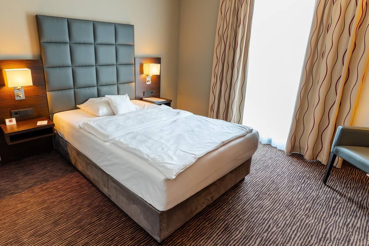 Une chambre d’hôtel avec un grand lit, deux tables de chevet avec des lampes, une tête de lit, un banc et une moquette à motifs.