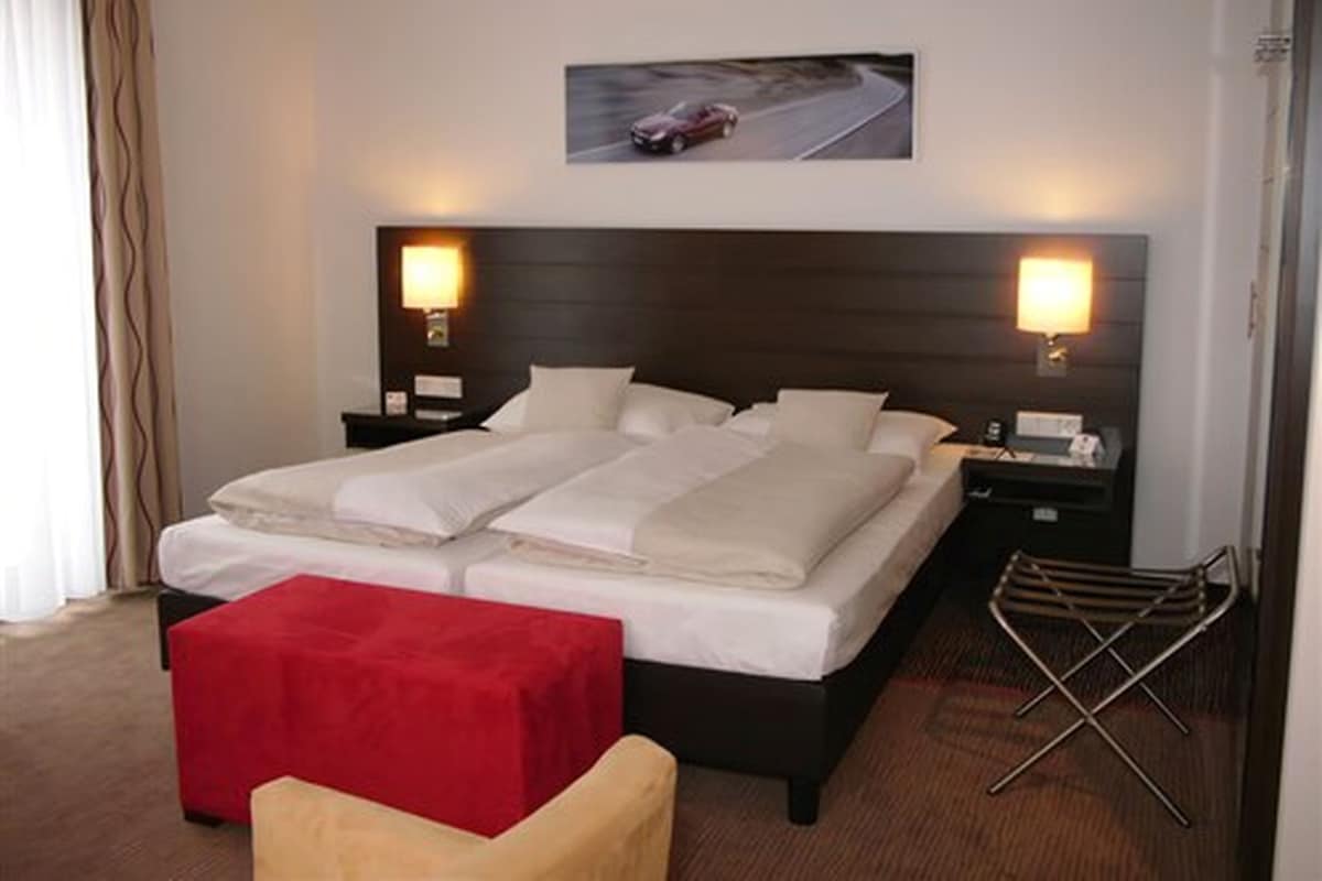 Quarto de hotel com uma cama grande, duas mesas de cabeceira com abajures, um banquinho vermelho e um quadro na parede de um carro em uma estrada.