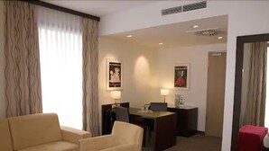 Ein Hotelzimmer mit Sofa, Ohrensessel und einem kleinen Tisch mit einem roten Gegenstand. Es gibt einen Schreibtisch mit Stuhl und Lampe.