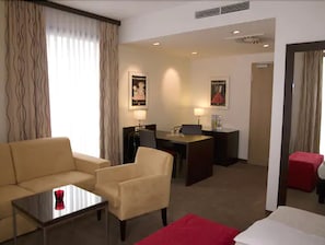 Quarto de hotel com sofá, poltrona e uma pequena mesa com um objeto vermelho. Há uma escrivaninha com cadeira e abajur.