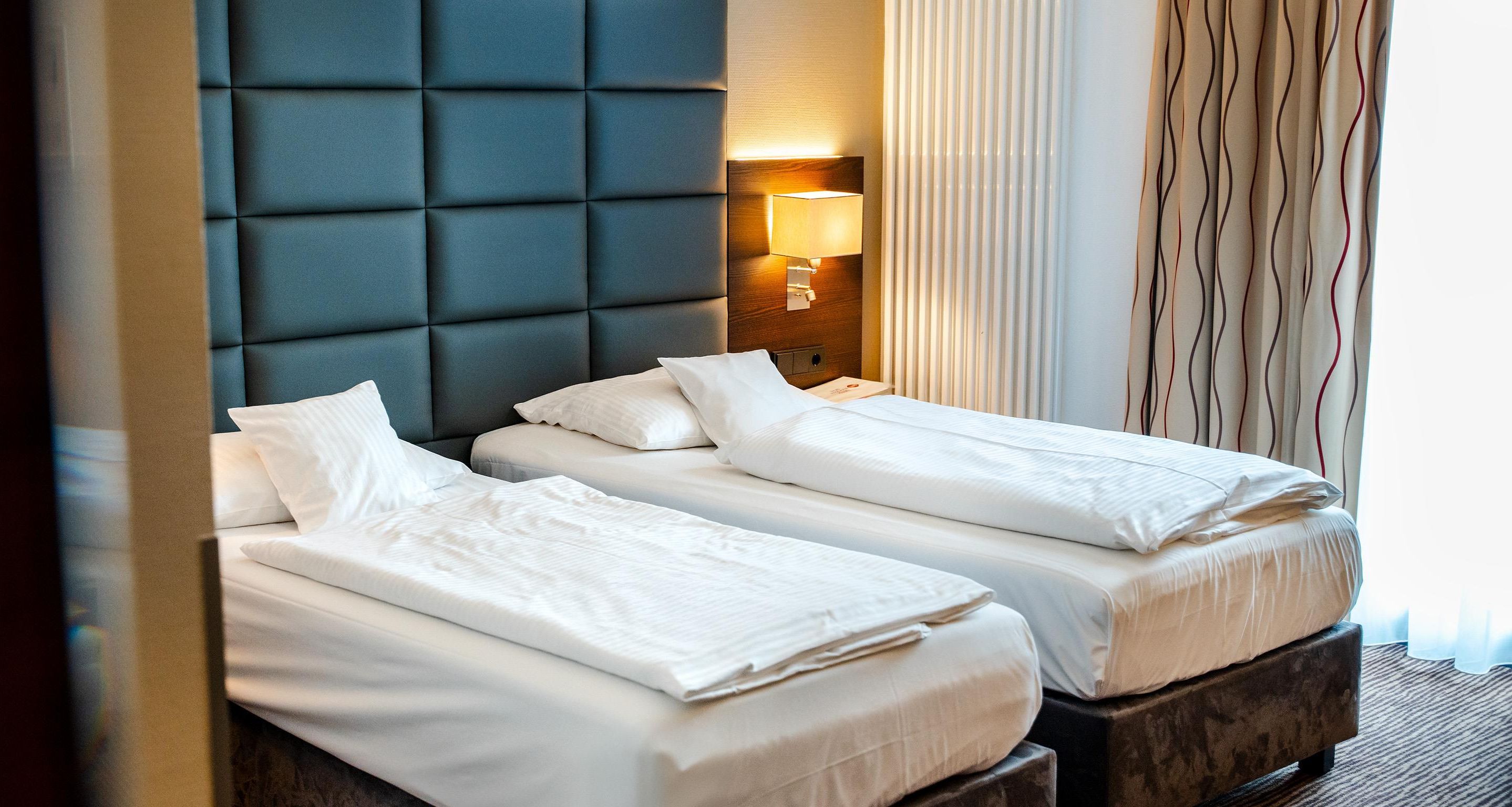 A hotel room with two beds, a padded headboard, a bedside lamp, and a window with curtains.