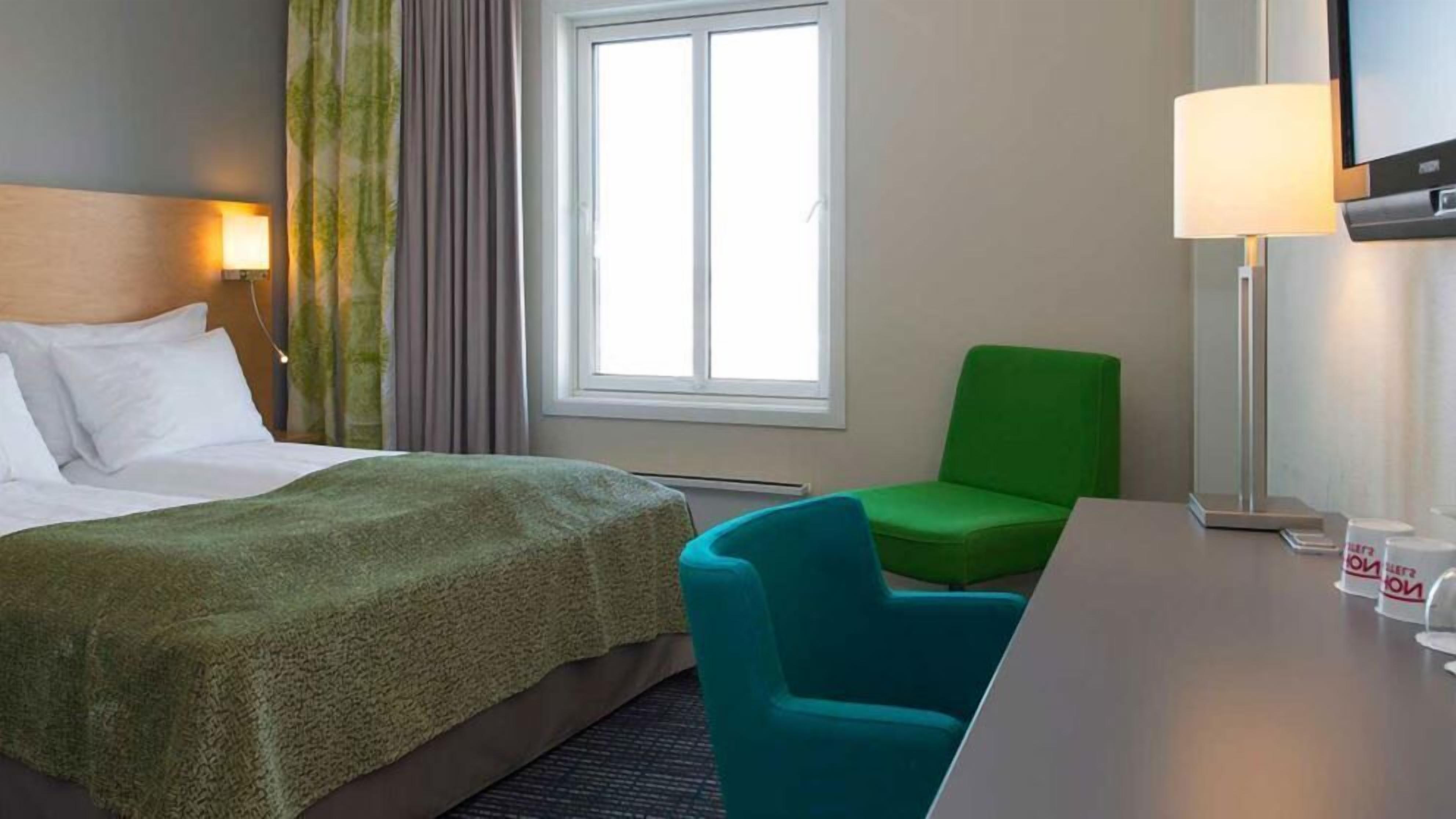 Ein Hotelzimmer mit einem Bett, einem grünen Sessel, einem Schreibtisch und einem Fernseher.