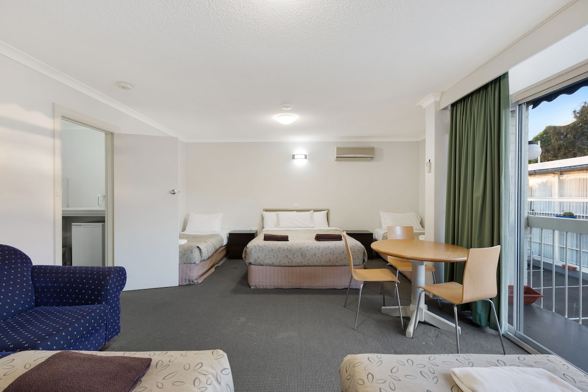 A hotel room with a bed, two bedside tables, a chair, a small table, and a view of the outside through a sliding glass door.
