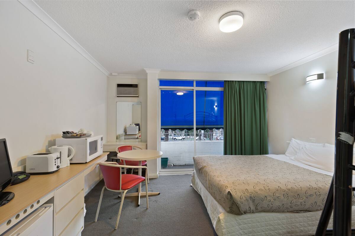 A hotel room with a bed, a desk with a computer, a chair, a microwave, and a window with a view of the outside.