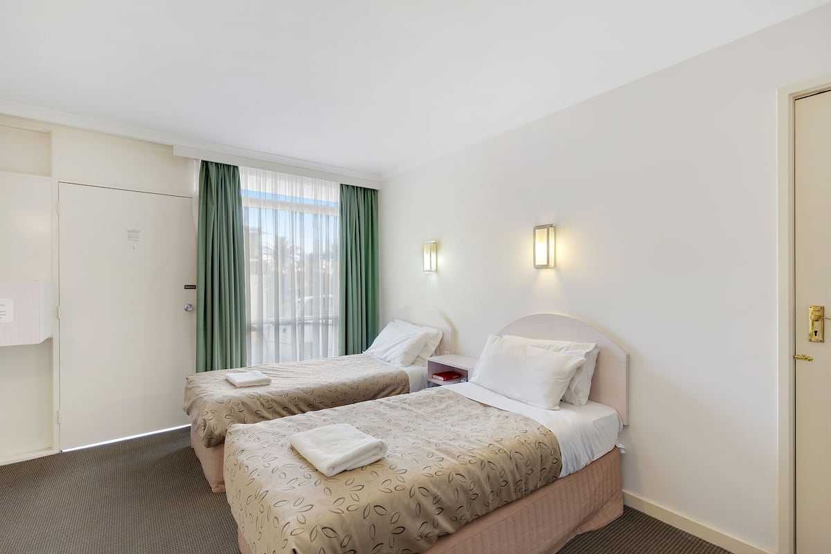 A hotel room with two beds, a window with green curtains, bedside lamps, and a door.