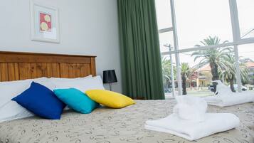 A hotel room with a bed, pillows, a window with curtains, a framed picture on the wall, and a view of palm trees and buildings outside.