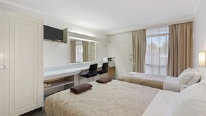 A hotel room with two beds, a desk with a chair, a TV mounted above the desk, and a window with curtains.
