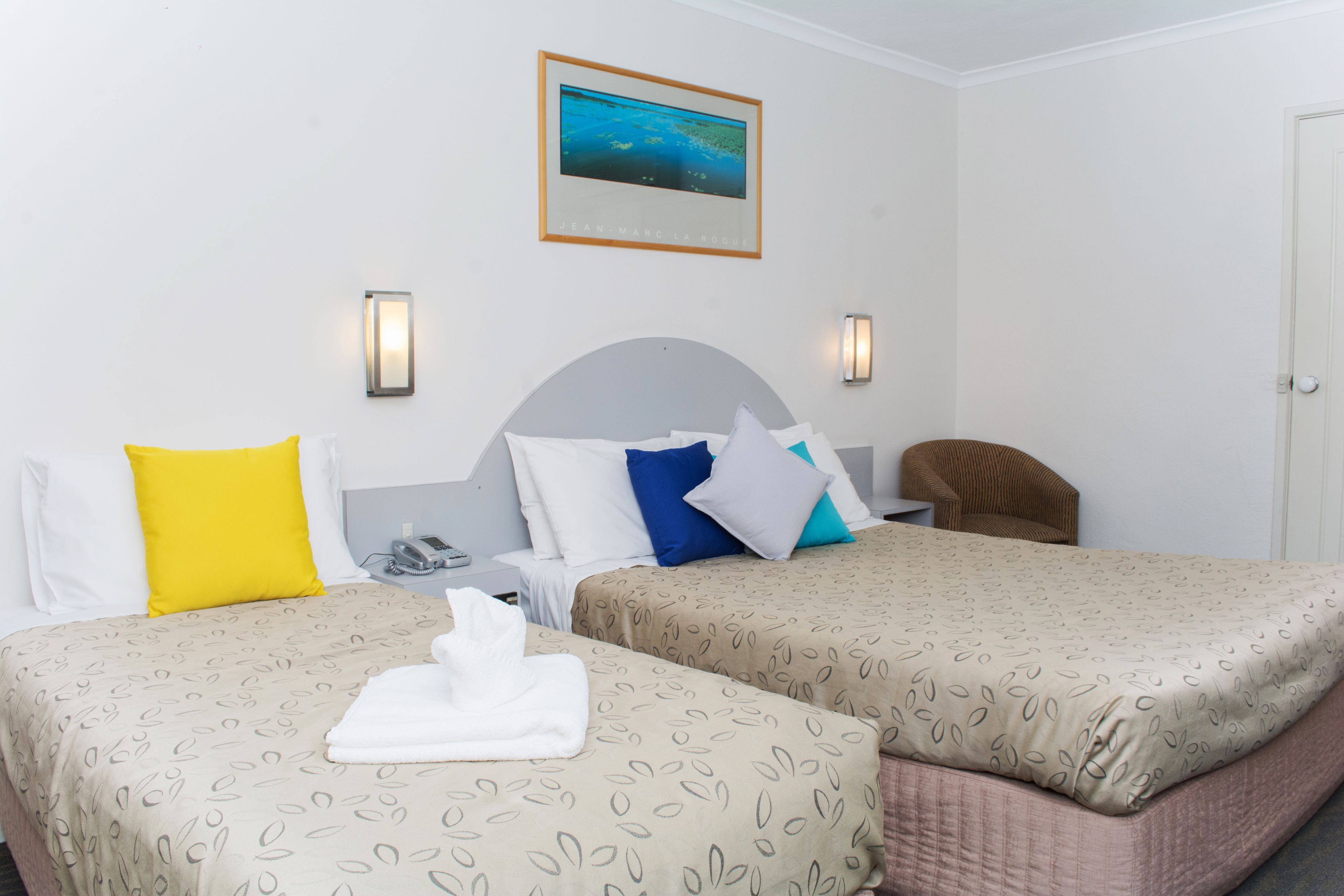 Una habitación de hotel con dos camas, una silla, un cuadro en la pared y un teléfono en la mesita de noche.