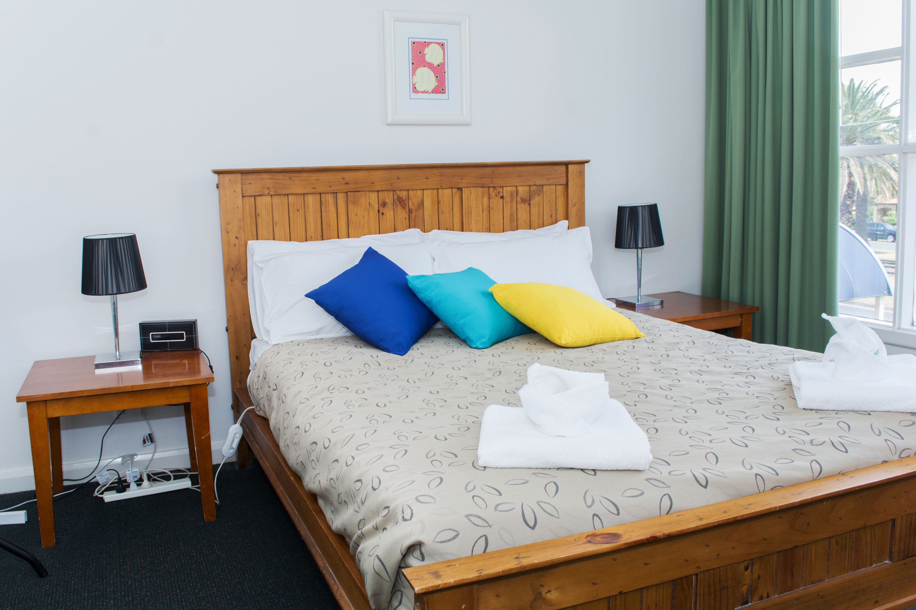 Een net opgemaakt bed met een beddegoed met dessin, twee nachtkastjes met lampen, een stopcontact en een ingelijst beeld aan de muur.