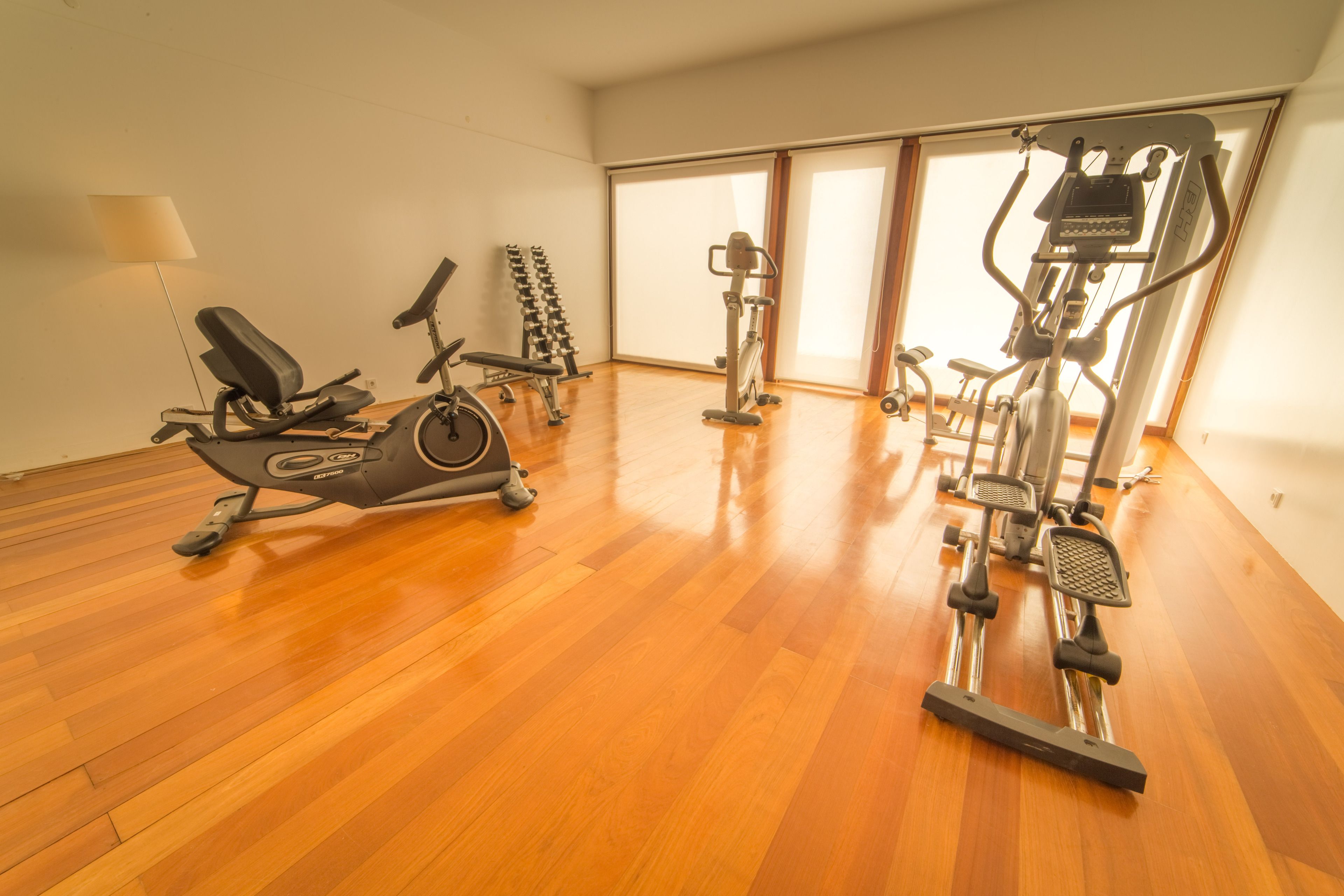 A gym with various exercise equipment, including an elliptical, a stationary bike, and a stair climber, on a wooden floor with large windows.
