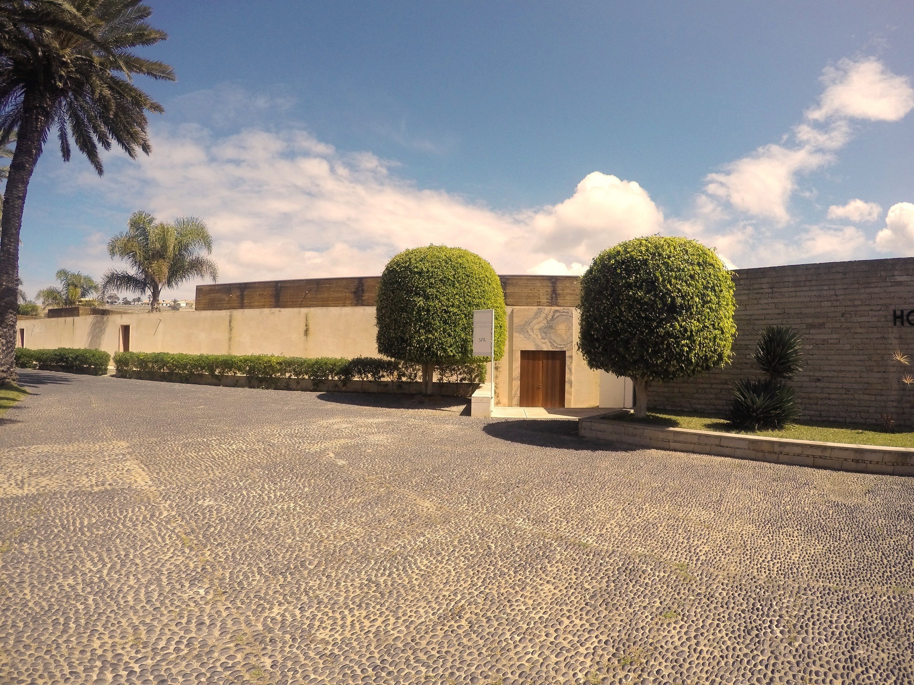 Área de entrada pavimentada com placa de spa, porta de madeira e parede de tijolos, com vista parcial de um edifício.
