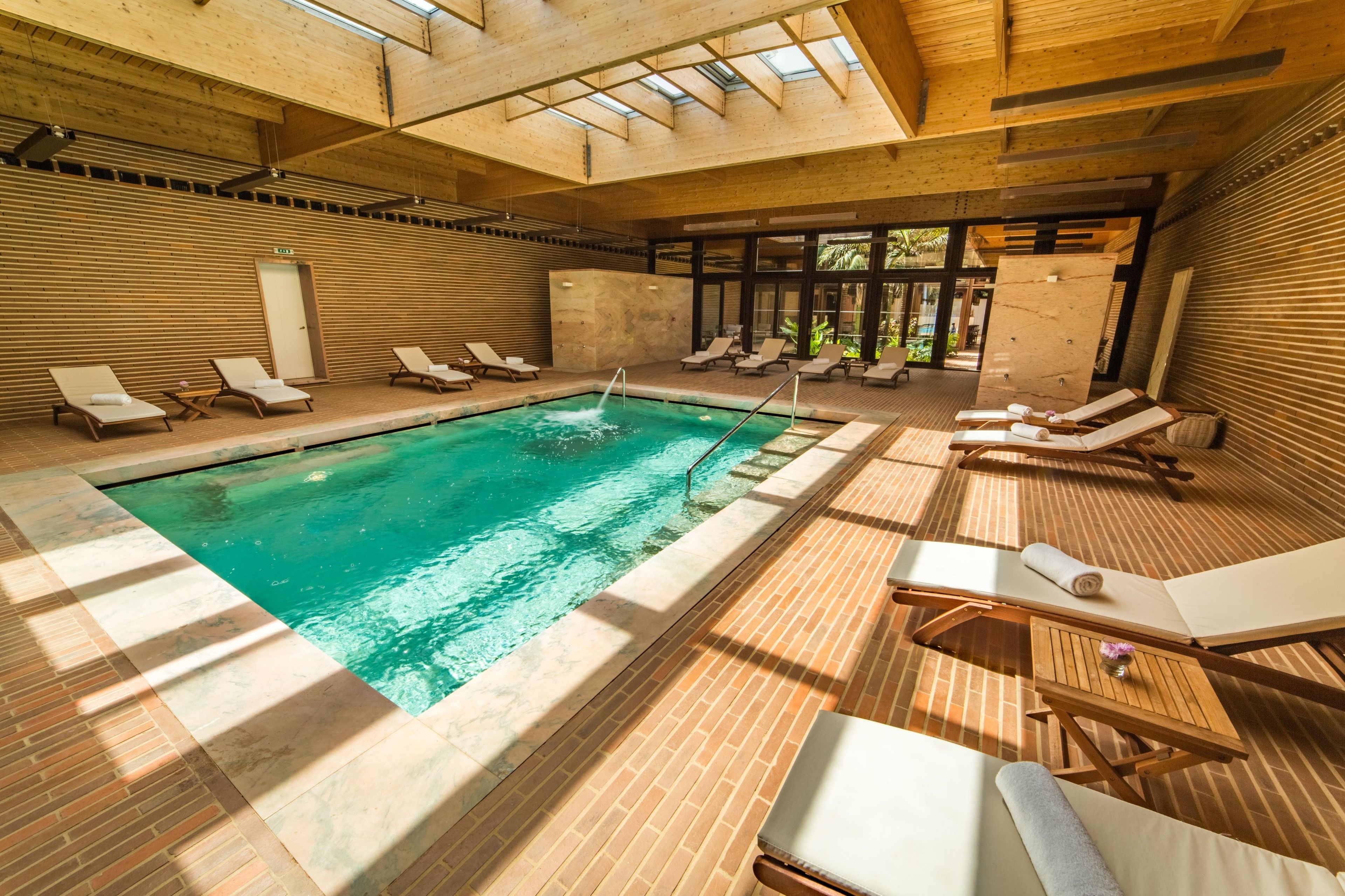 A modern indoor swimming pool with wooden flooring, sunlit by skylights, surrounded by lounge chairs and a small table with a vase.