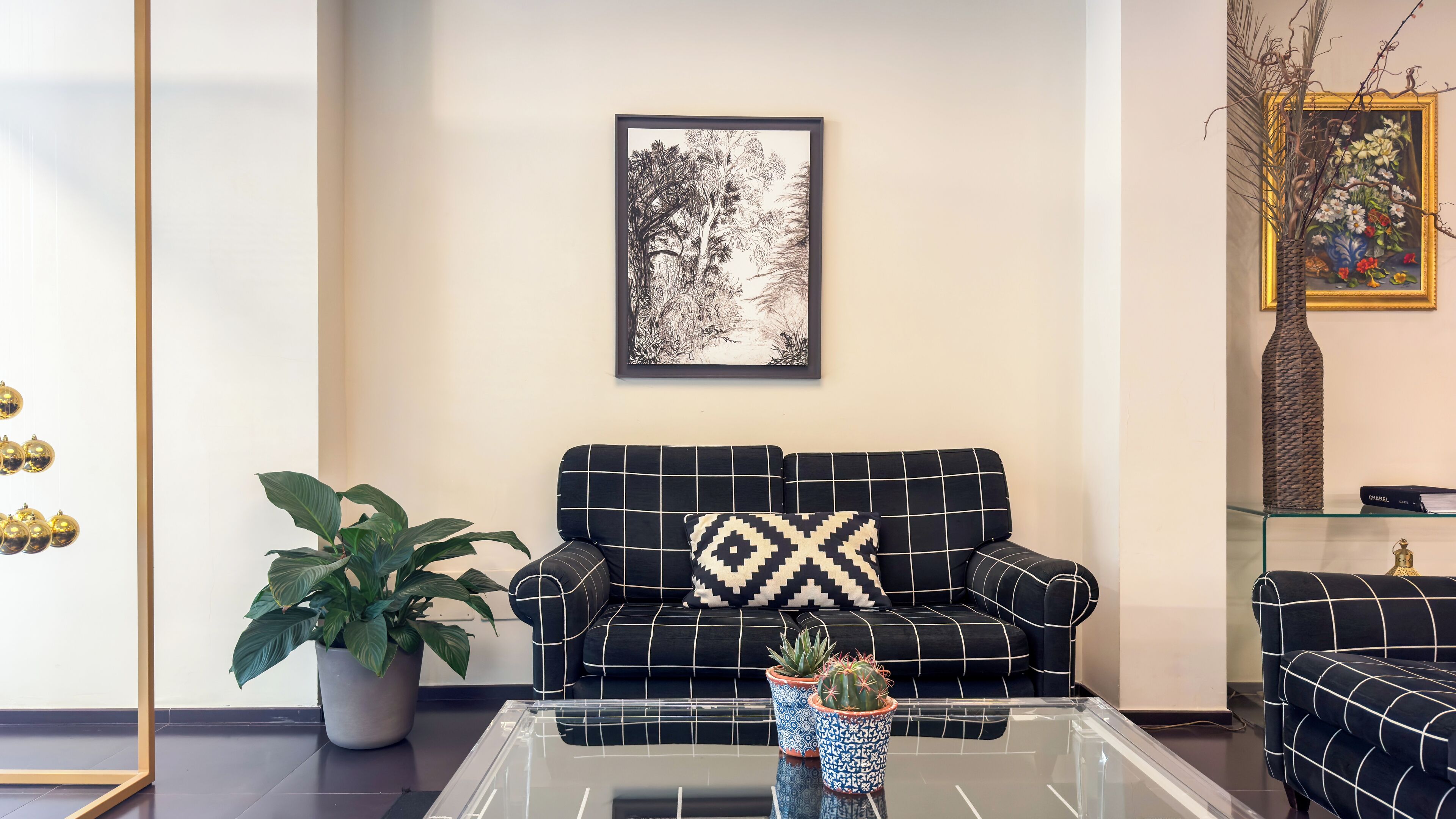 Una sala de estar con un sofá a cuadros blanco y negro, una planta grande en maceta, una mesa de centro de cristal y una obra de arte enmarcada en la pared.
