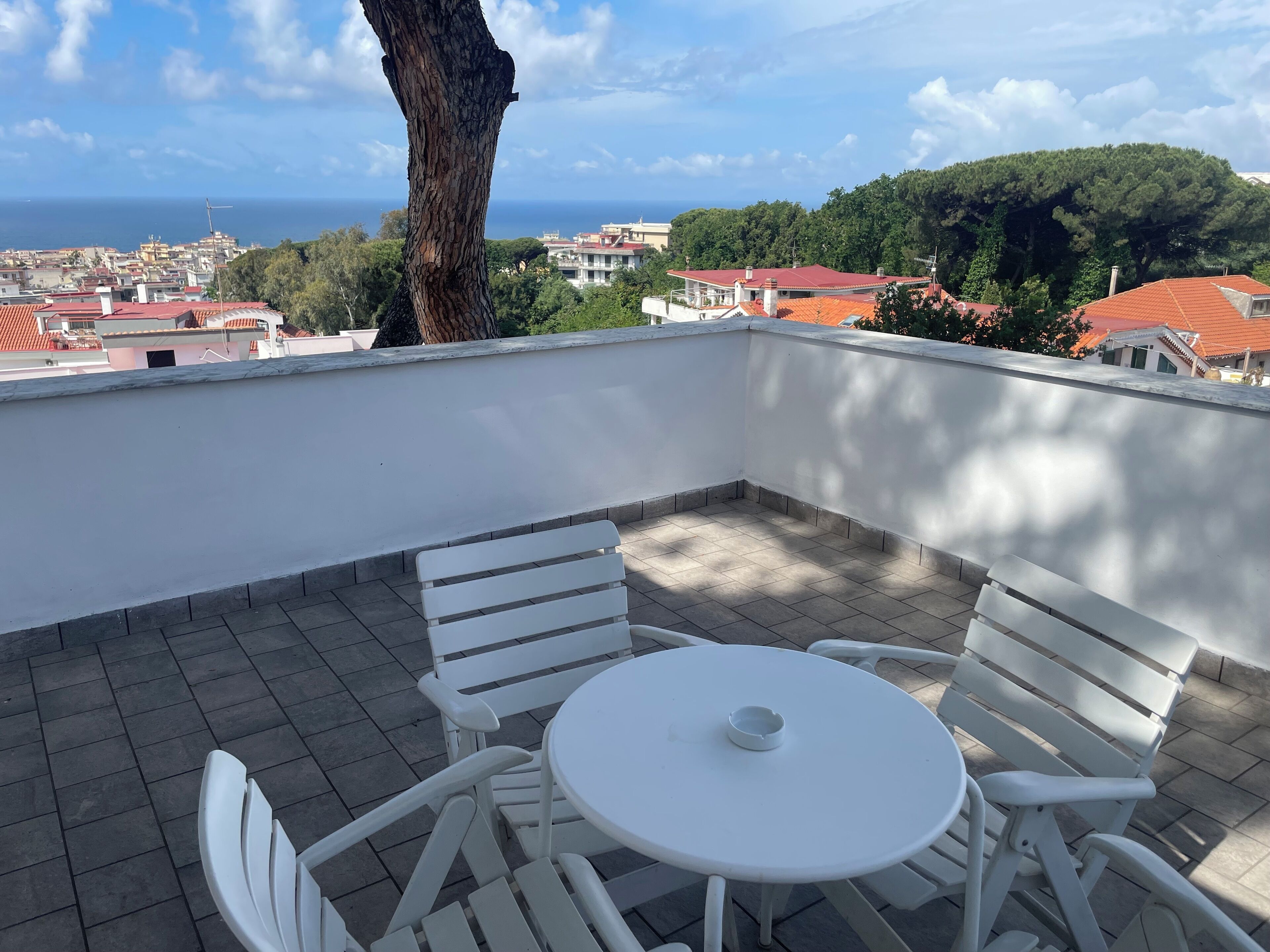 Une terrasse avec une table et des chaises blanches, donnant sur une ville côtière aux bâtiments aux toits rouges et offrant une vue sur la mer.