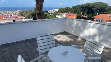 Une terrasse avec une table et des chaises blanches, donnant sur une ville côtière aux bâtiments aux toits rouges et offrant une vue sur la mer.
