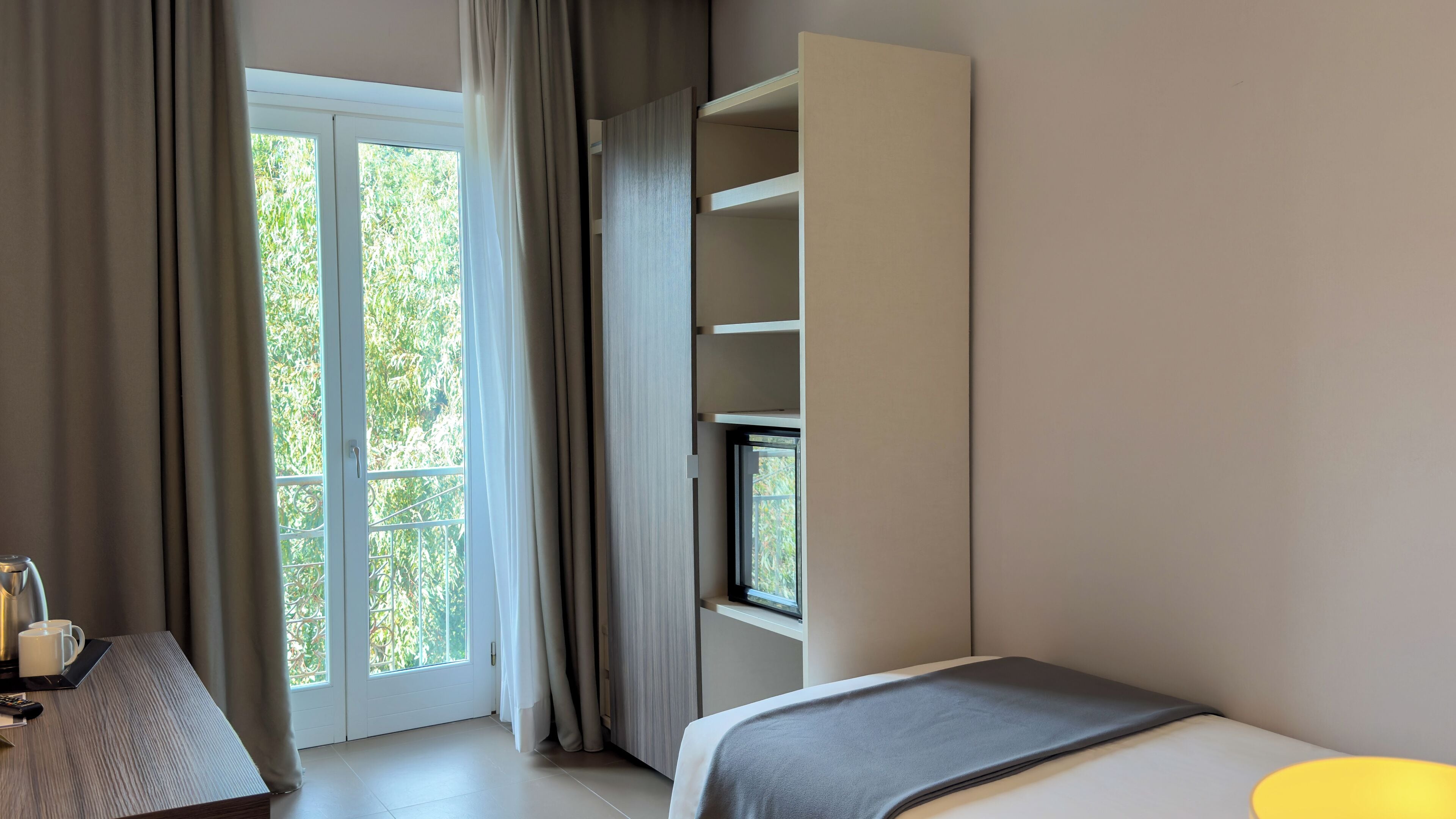 Una habitación de hotel moderna con un amplio ventanal corredero que da a un balcón, una cama con una manta gris y un mueble empotrado para la televisión.