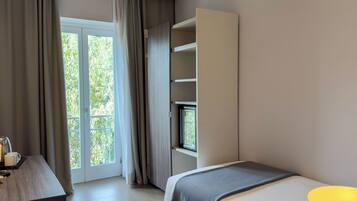 Una habitación de hotel moderna con un amplio ventanal corredero que da a un balcón, una cama con una manta gris y un mueble empotrado para la televisión.