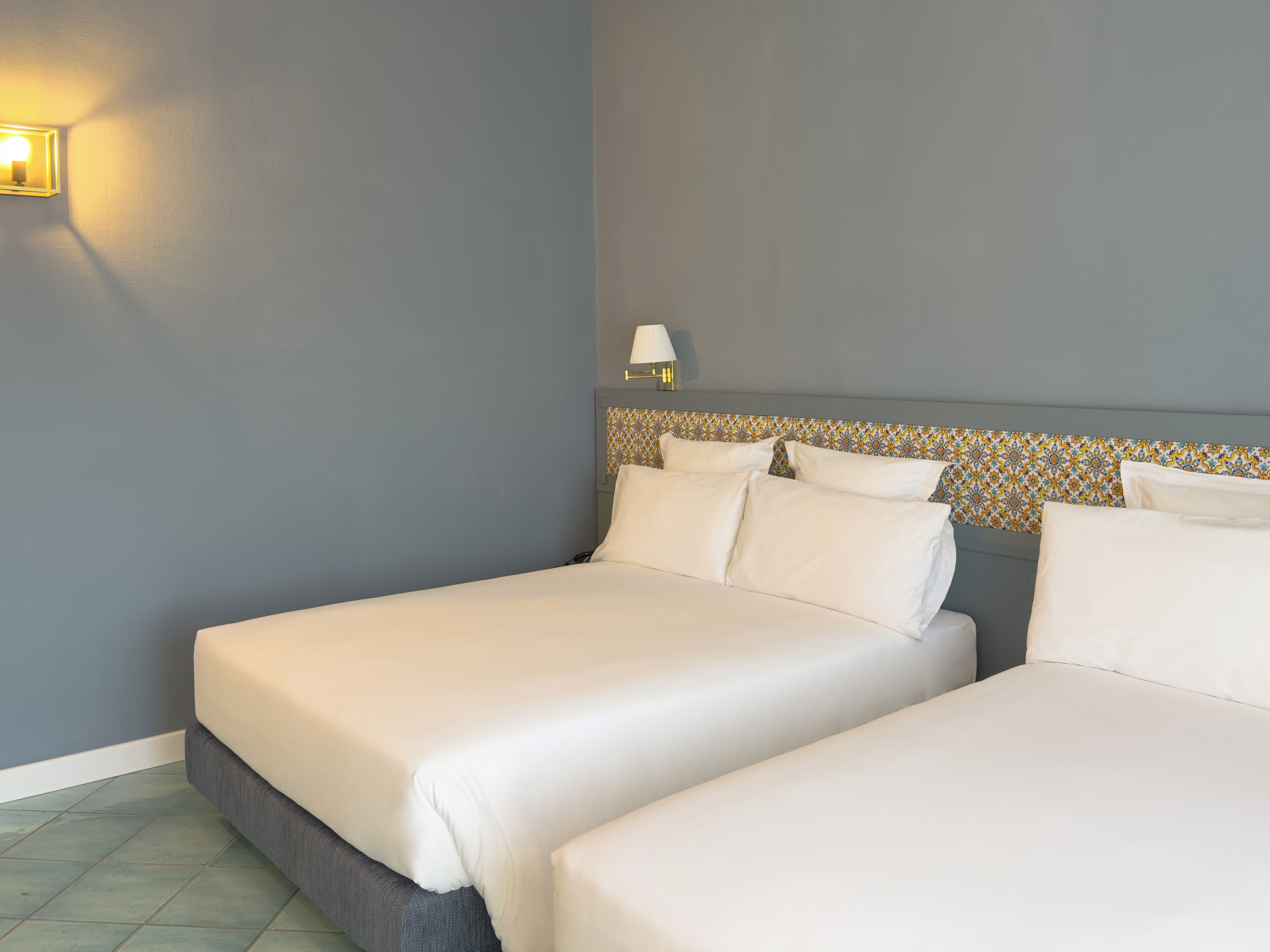 A hotel room with two beds, a patterned headboard, a wall-mounted lamp, and a tiled floor.
