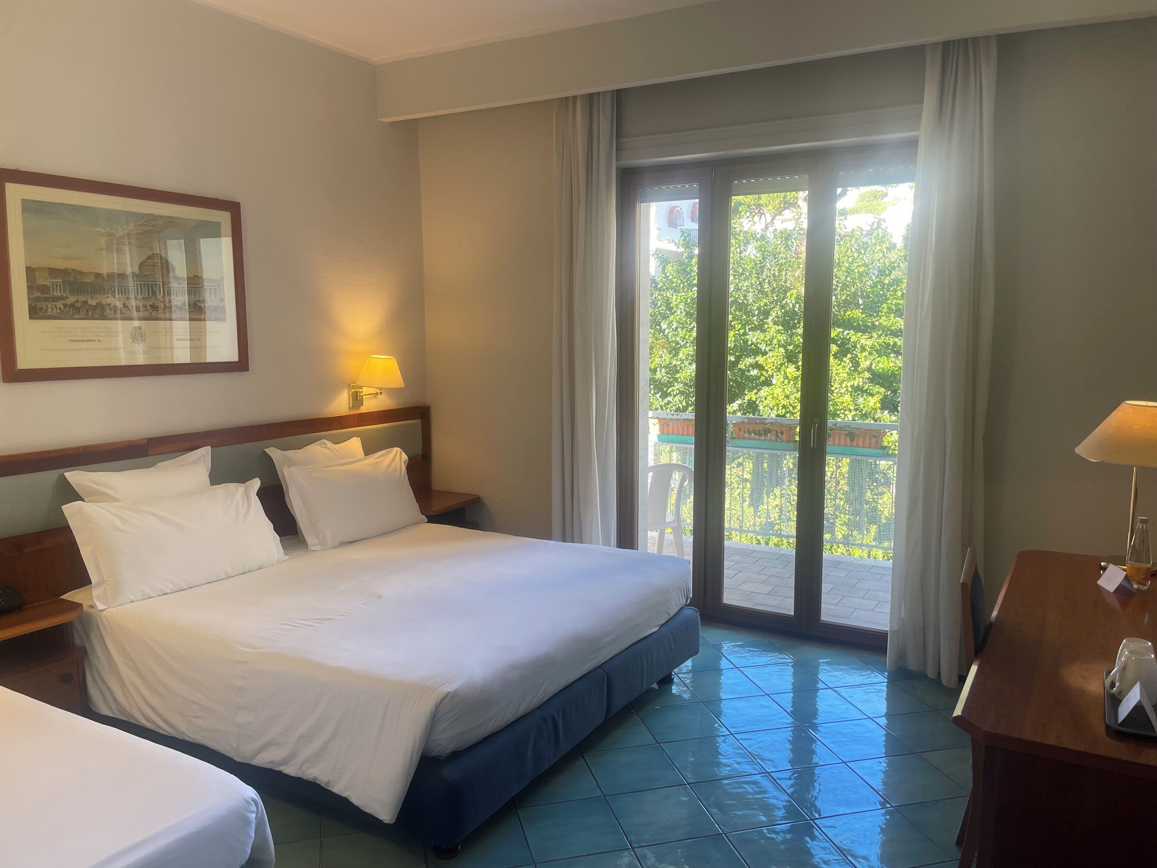 A hotel room with a bed, a desk, and a balcony with a view of greenery.