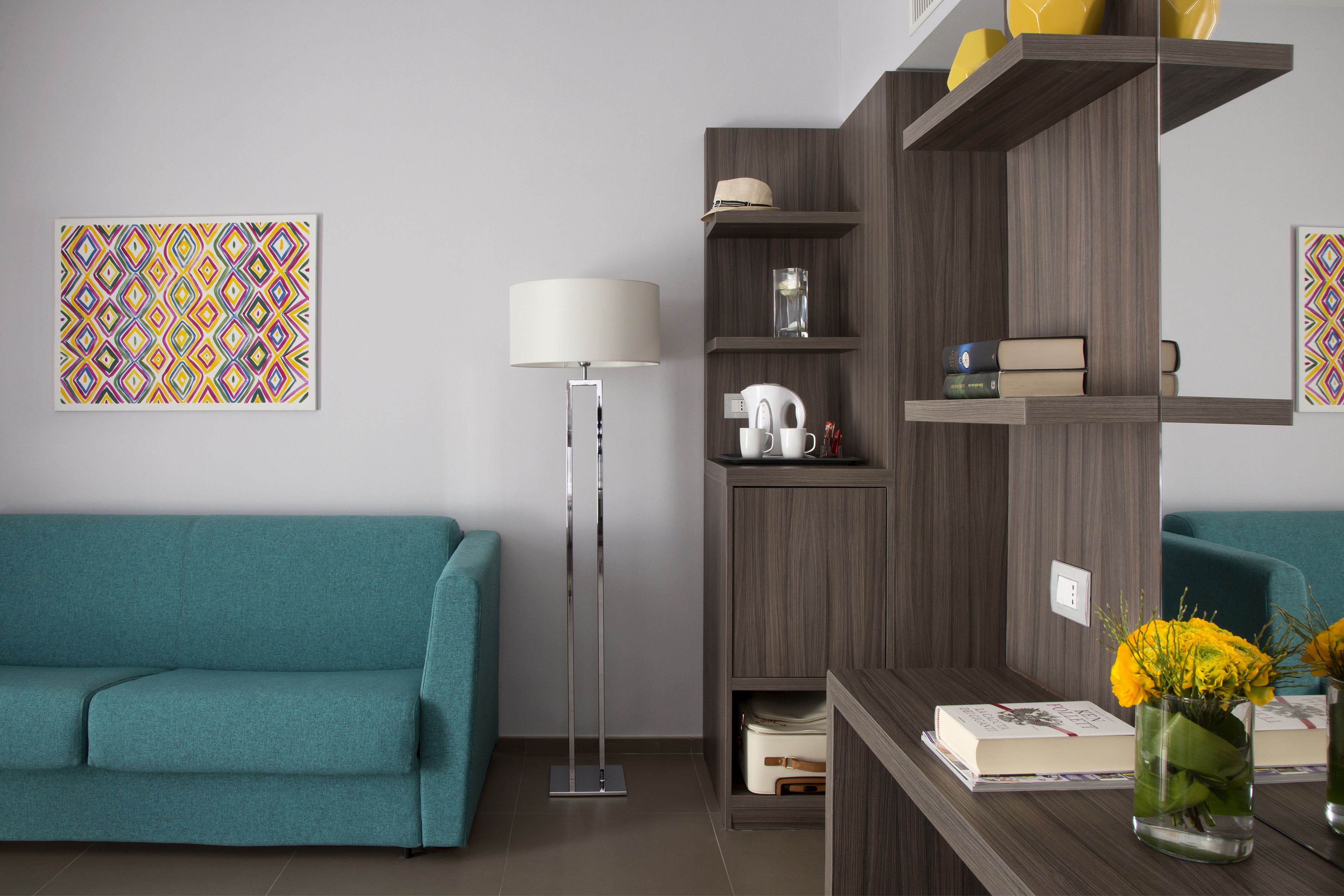 A modern living room with a teal sofa, a floor lamp, a wooden bookshelf, and a decorative vase with flowers.