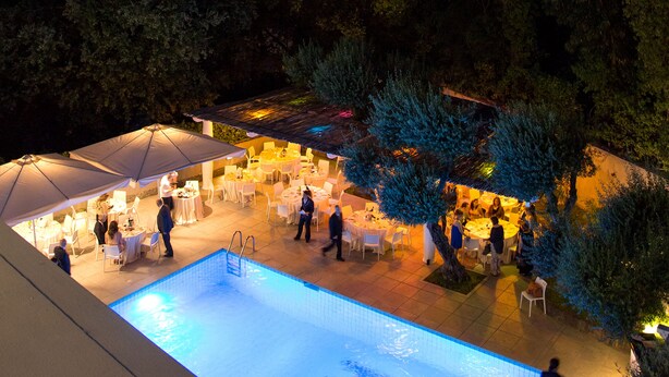 Zona de piscina exterior con mesas y sillas, rodeada de árboles y un cielo despejado.