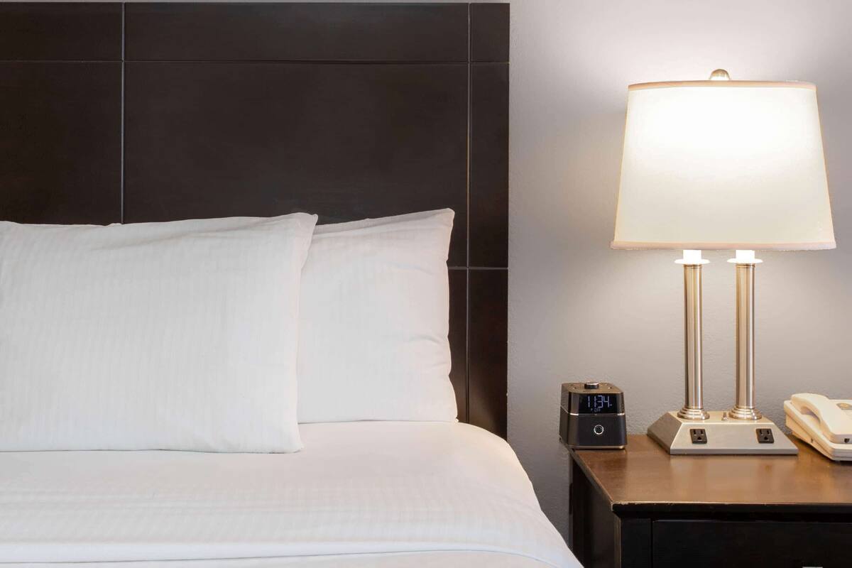 Un lit bien fait, avec du linge de lit blanc, une tête de lit sombre, une table de chevet avec une lampe, une horloge et un téléphone.