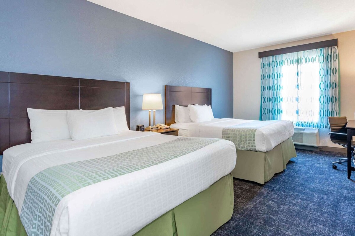 Habitación de hotel con dos camas, un escritorio, una silla, una lámpara y una ventana con cortinas.