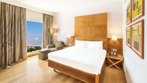 Habitación de hotel con una cama grande, un sofá, una mesita de noche con lámparas, vistas a la ciudad y una ventana con cortinas.