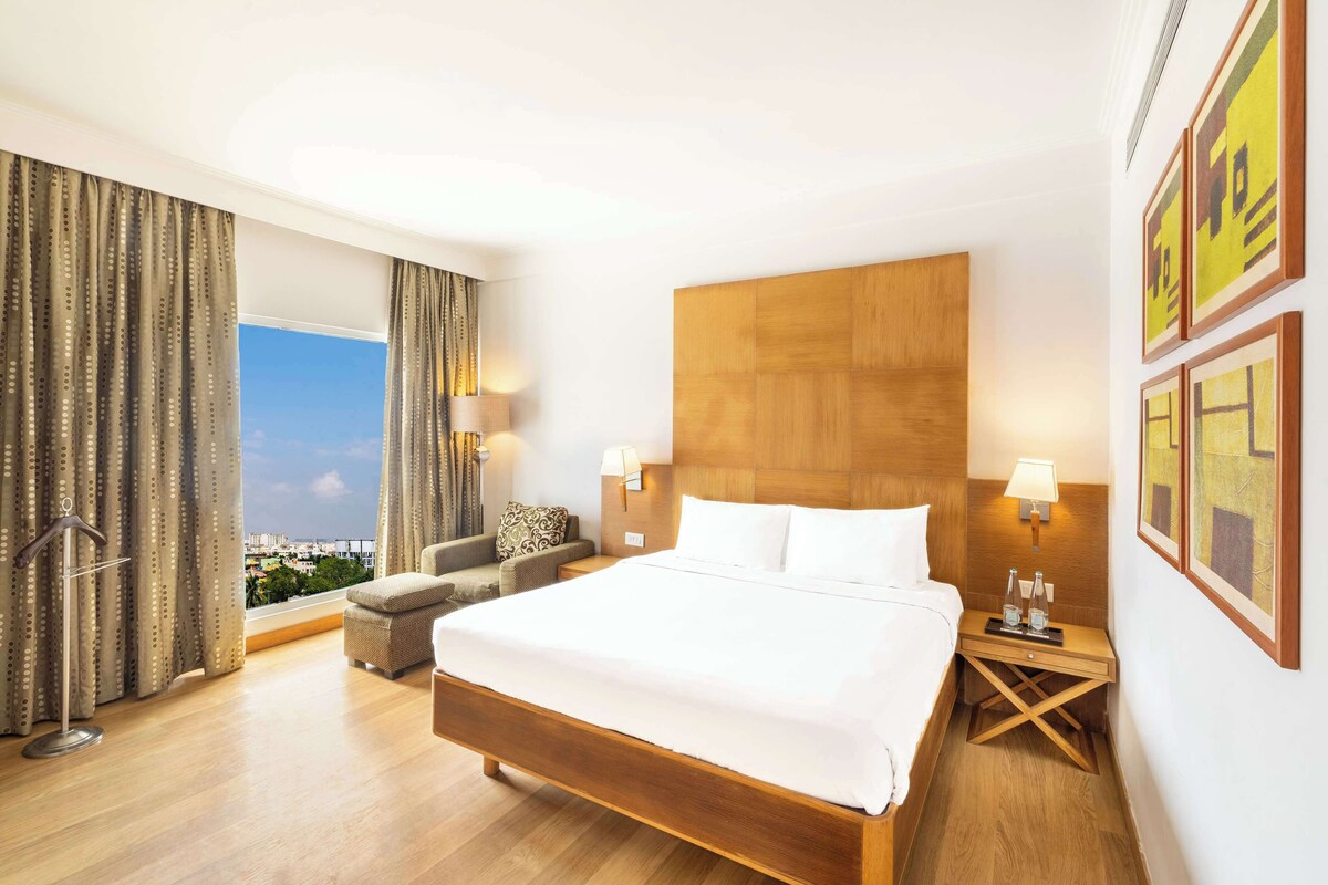 Habitación de hotel con una cama grande, un sofá, una mesita de noche con lámparas, vistas a la ciudad y una ventana con cortinas.