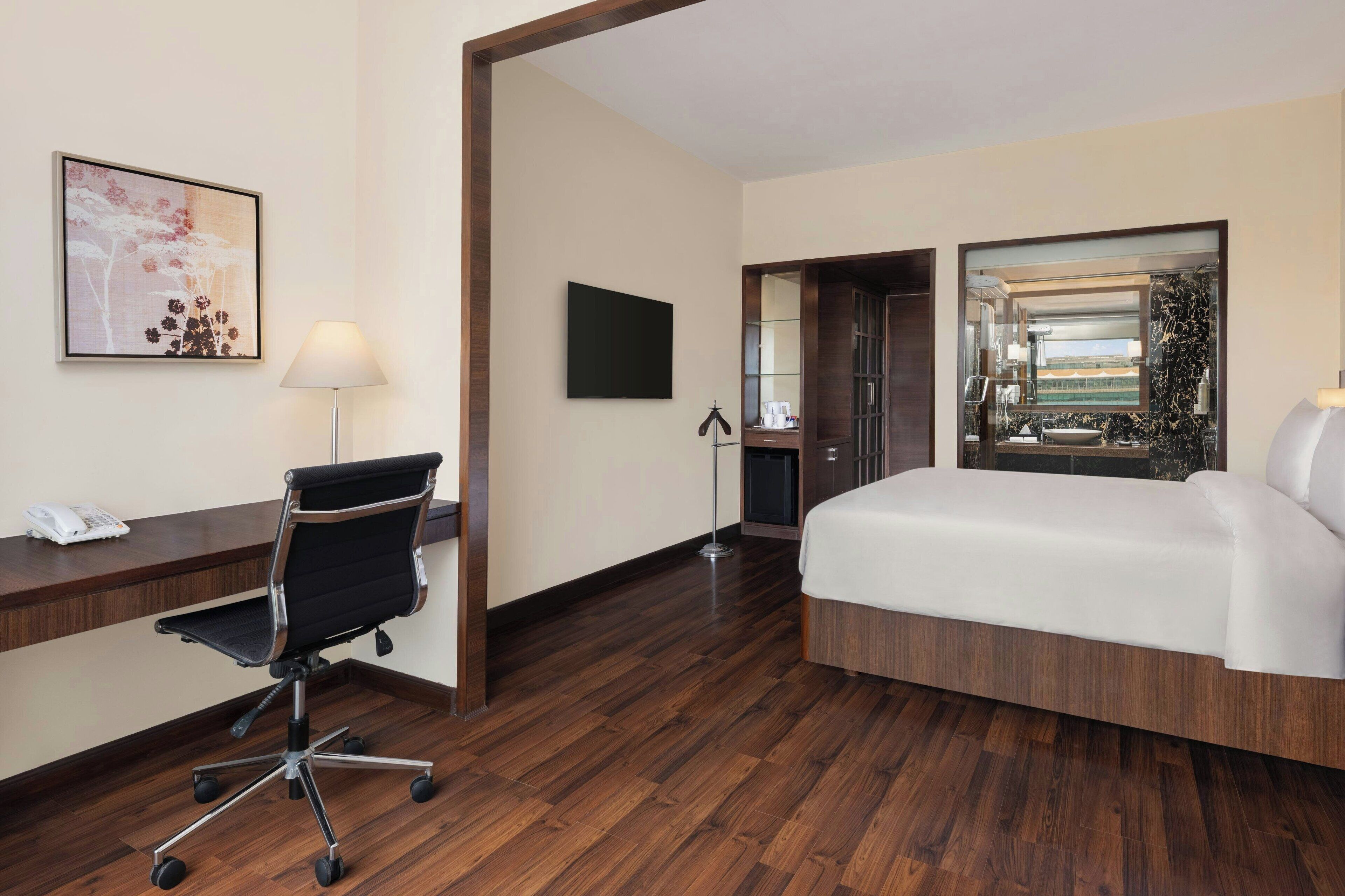 A hotel room with a wooden floor, a desk with a chair, a bed, a TV, and a bathroom visible through an open door.