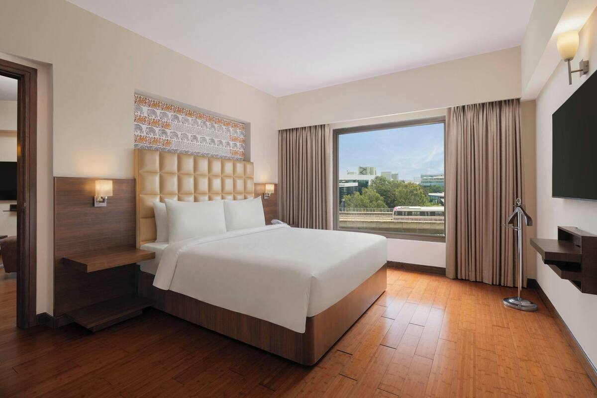 A modern hotel room with a large bed, a headboard with a patterned design, a bedside table with a lamp, a large window with curtains, and a flat-screen TV mounted on the wall.