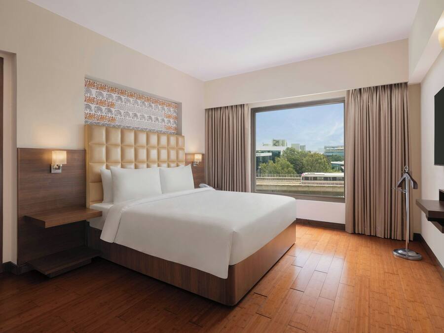 Una habitación de hotel moderna con una cama grande, cabecera con diseño estampado, mesita de noche con lámpara, ventana amplia con cortinas y televisor de pantalla plana montado en la pared.