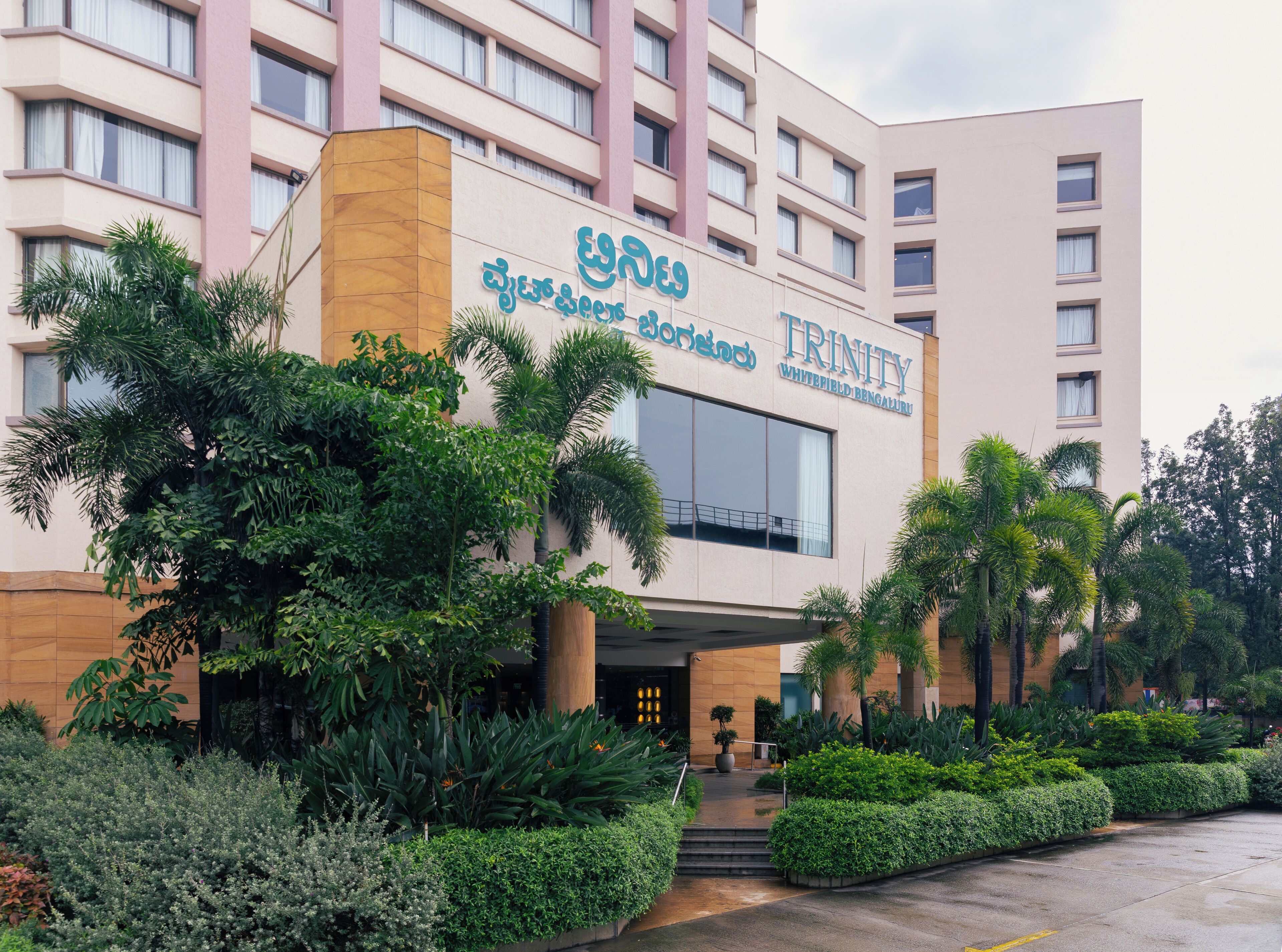 Un hôtel appelé Trinity Whitefield Bangalore, doté d’une architecture moderne, se distingue par son entrée imposante et la verdure luxuriante qui l’entoure.
