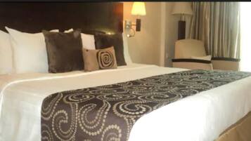 A hotel room with a large bed, a headboard, a lamp, a chair, and a window with curtains.