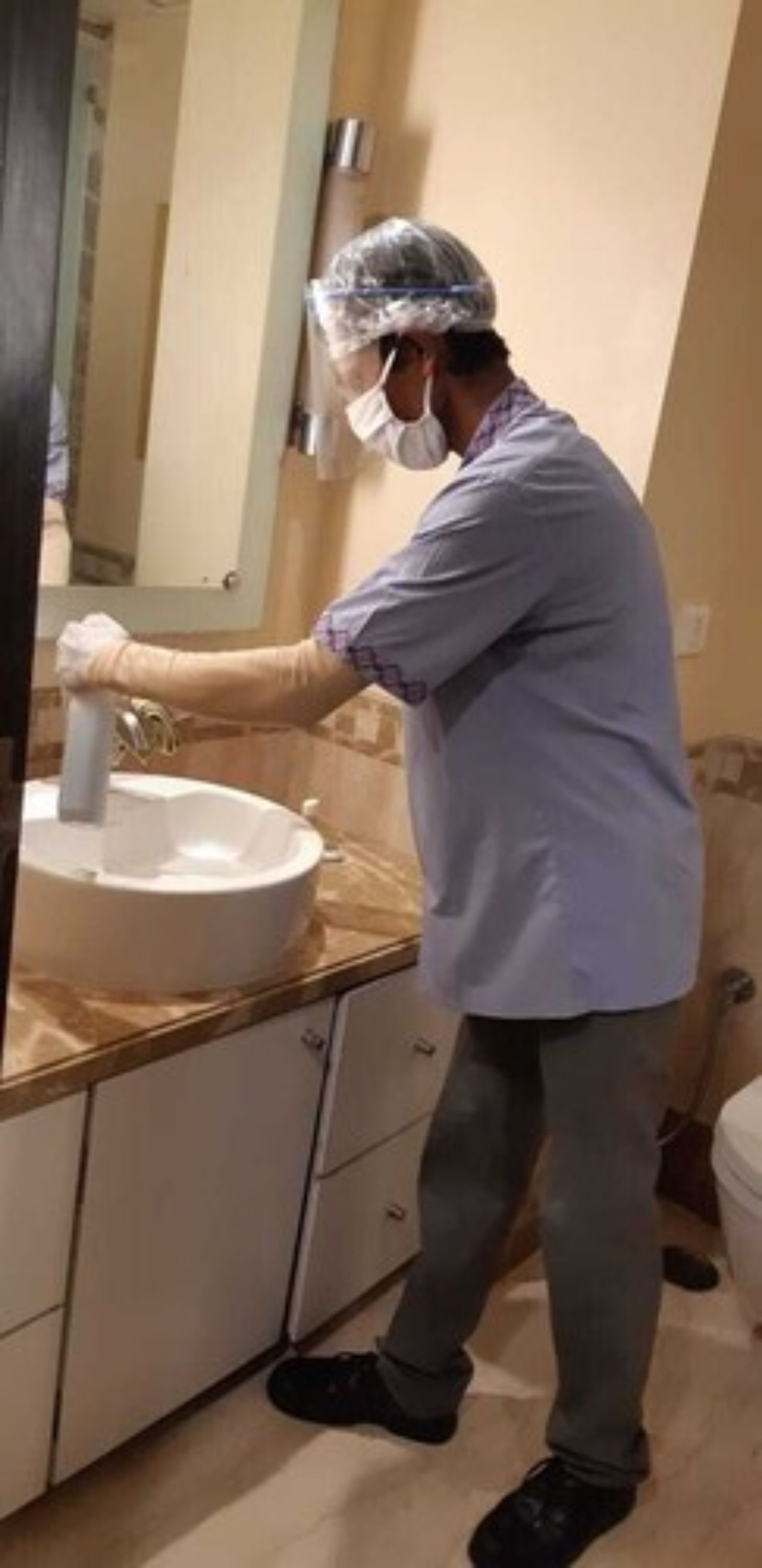 A person in a blue shirt and protective gear working on a sink in a bathroom with beige walls and a wooden countertop.