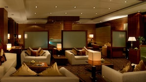 Un vestíbulo de hotel con varias zonas de estar, incluyendo sofás y sillones, dispuestos alrededor de una mesa de centro. Varias lámparas proporcionan iluminación, y la habitación cuenta con un espejo grande y paneles de madera en las paredes.