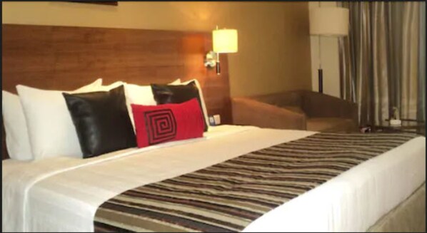 Una habitación de hotel con una cama grande, un cabecero de madera, almohada roja y negra, alfombra a rayas y una silla.