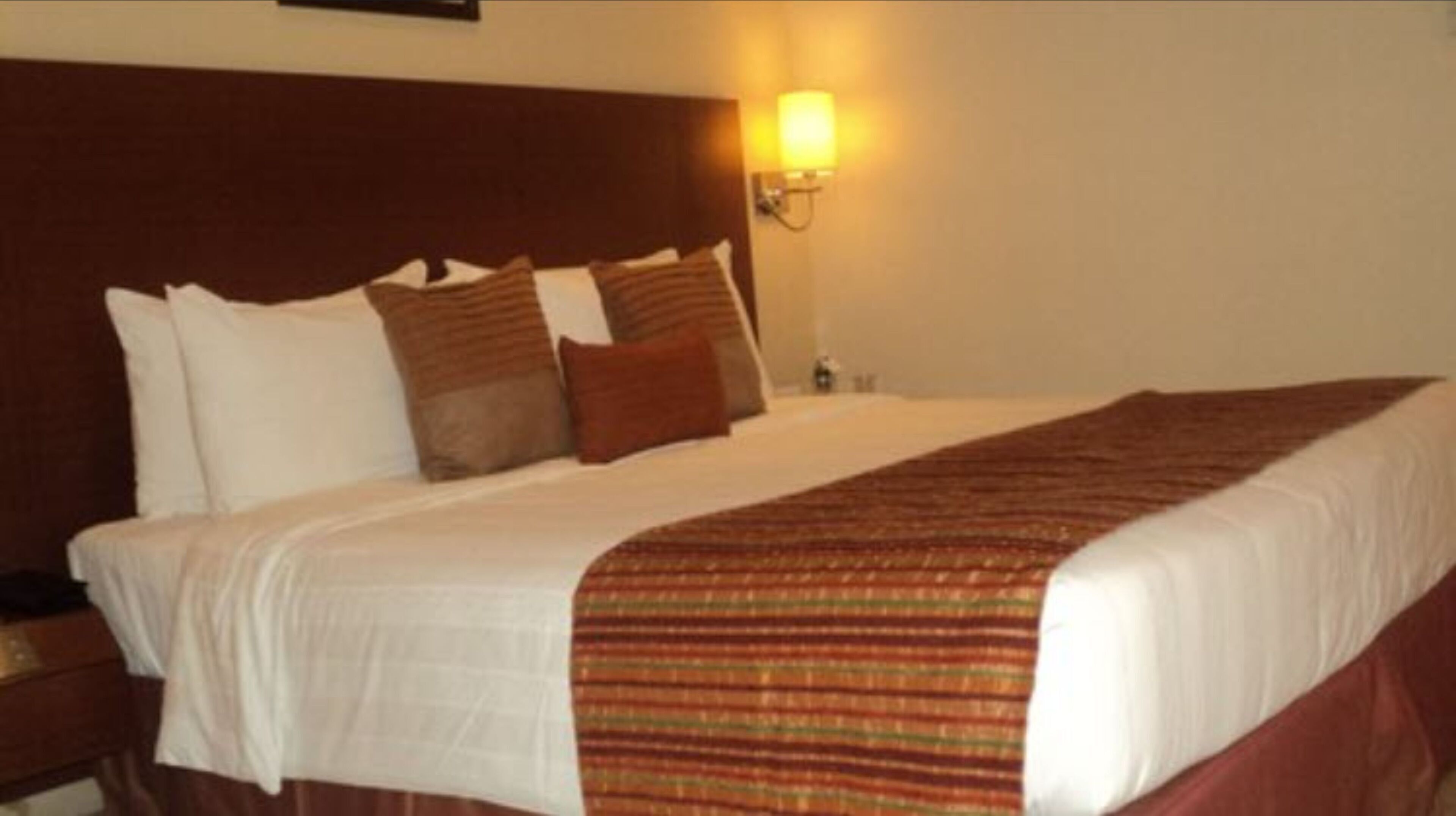 A neatly made bed with white and brown bedding, a striped blanket, and a wall-mounted lamp.