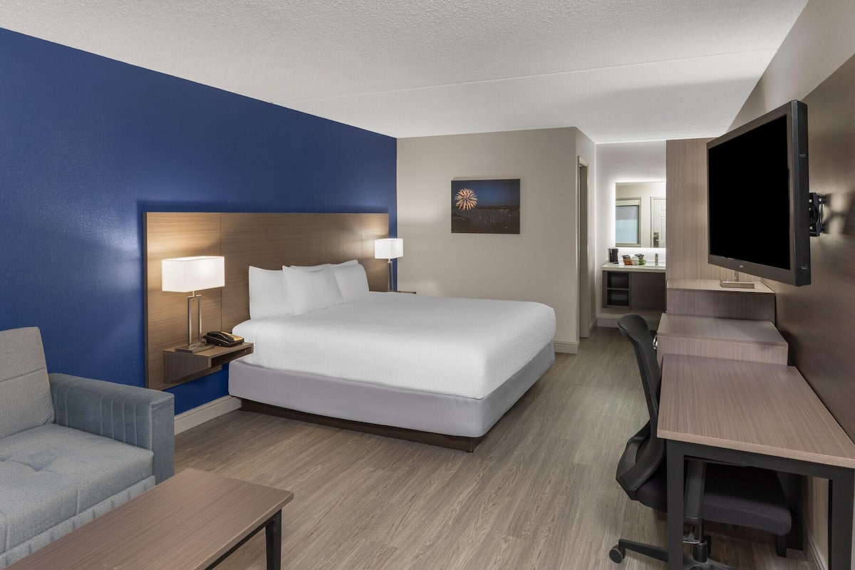 A hotel room with a bed, bedside tables, a desk, a chair, a TV, and a bathroom visible in the background.