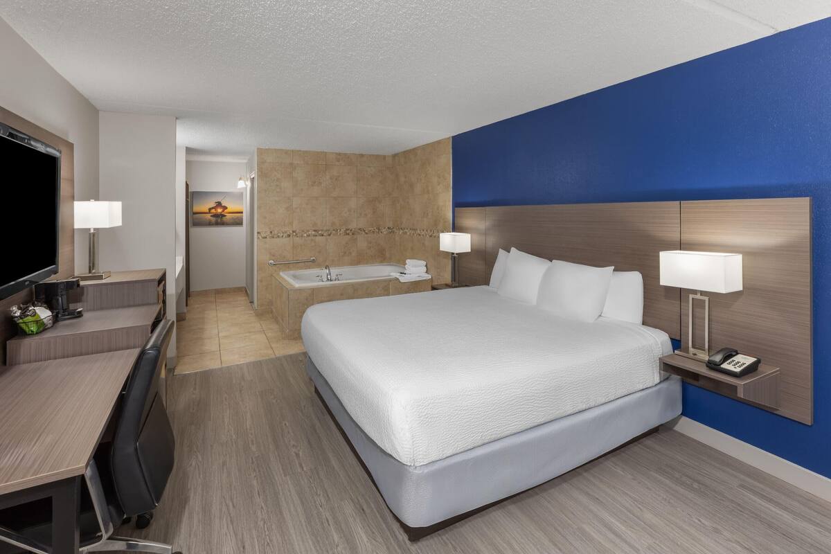 A hotel room with a bed, desk, chair, lamp, and a view of a sunset through the window.