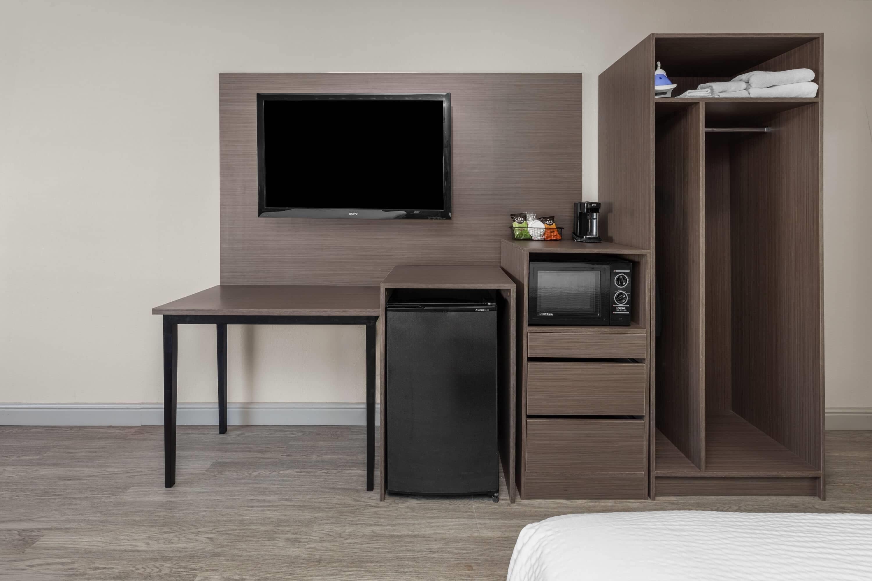 A hotel room with a flat-screen TV mounted on the wall, a small refrigerator, a microwave, a desk, and a wardrobe.
